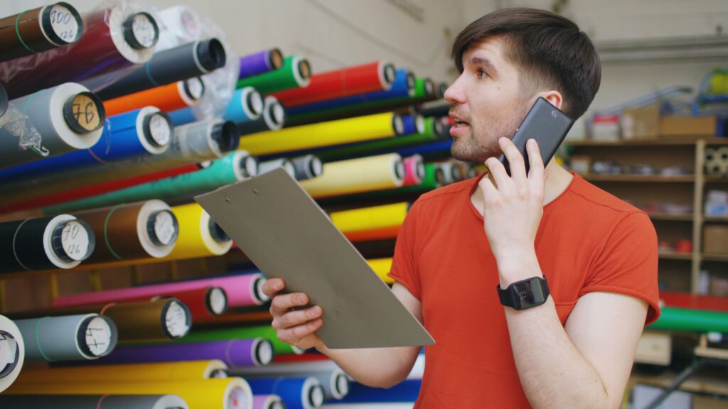 Man talking on phone with clipboard in vibrant textile store, organized fabric rolls in background.